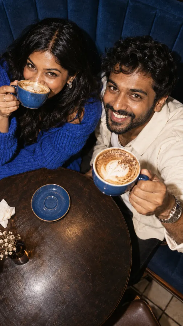AI-created overhead photo of two fictional Tamil adults playfully raising blue coffee cups toward the camera.